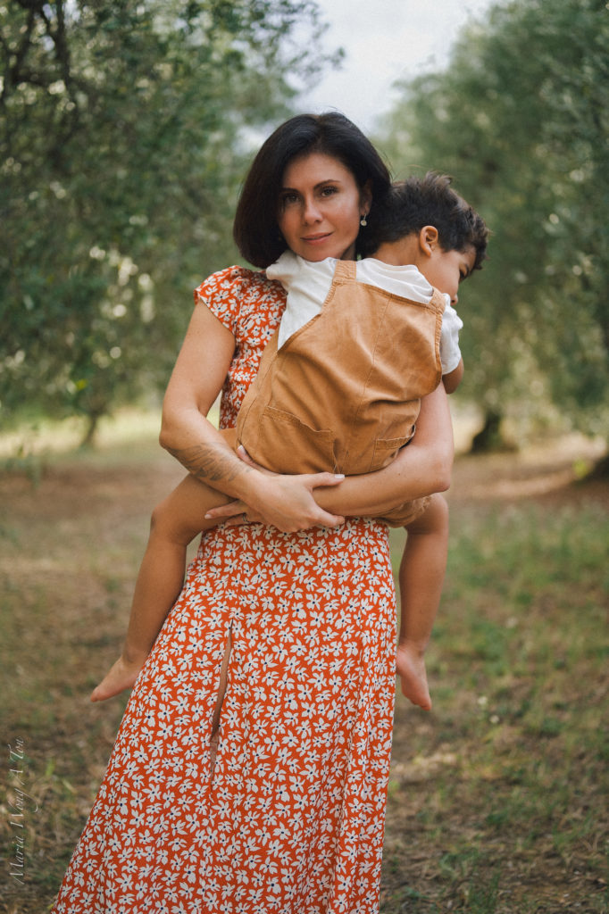 Portrait of a serene woman in a red floral dress holding a sleeping toddler in a tan carrier against her hip, standing amidst an orchard with soft-focus greenery in the background, depicting a peaceful moment of motherly care.