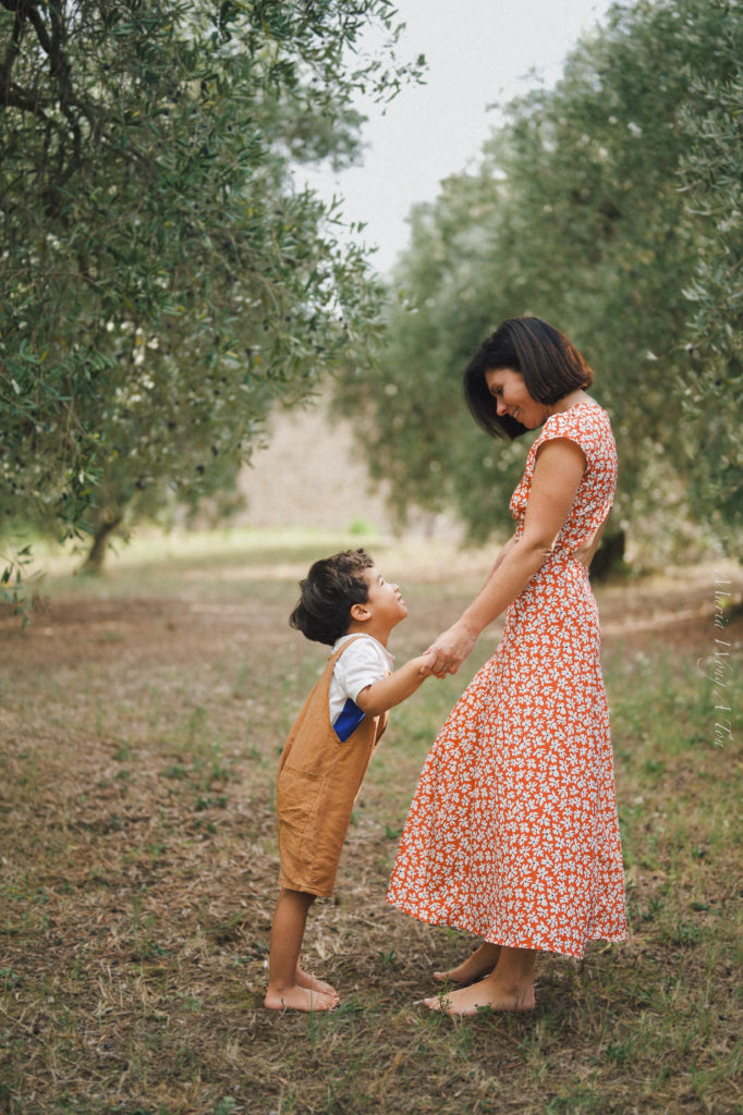 Portrait of a serene woman in a red floral dress holding a toddler, standing amidst an orchard with soft-focus greenery in the background, depicting a peaceful moment of motherly care.