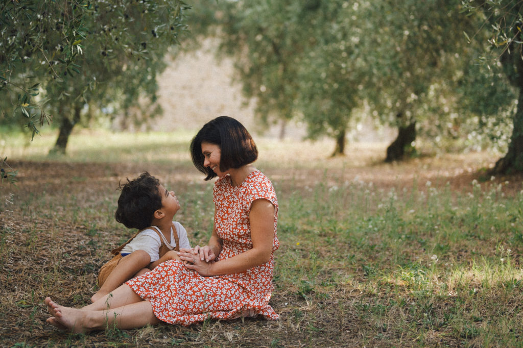 Smiling woman in a red floral dress seated on the ground in an olive grove, interacting joyfully with a young child who looks up at her; they touch hands, expressing a warm, familial connection, with trees softly blurred in the background.