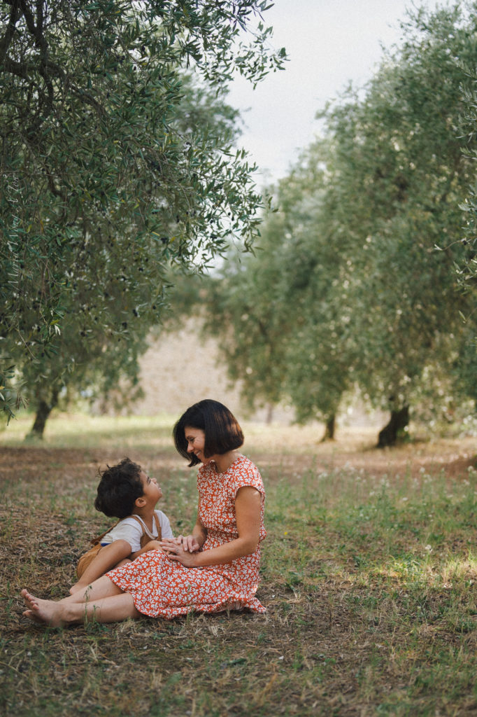 Smiling woman in a red floral dress seated on the ground in an olive grove, interacting joyfully with a young child who looks up at her; they touch hands, expressing a warm, familial connection, with trees softly blurred in the background.