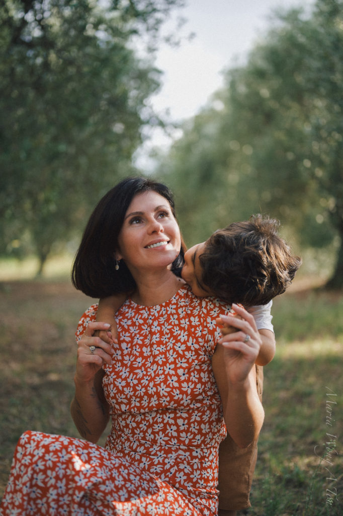 A joyful woman in a red floral dress looks upward, smiling as she receives a sweet kiss on the cheek from a small child, whose back is to the camera. They are surrounded by the soft, natural blur of an orchard, creating an atmosphere of intimate connection and love.