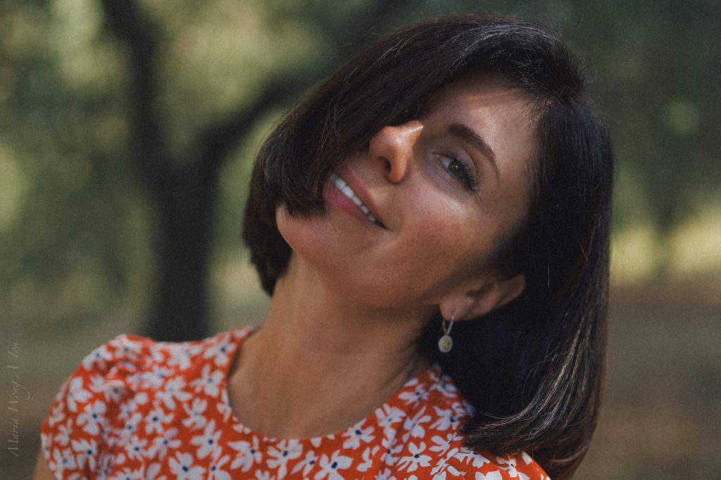 Close-up of a woman with a content smile, looking off to the side, set against a blurred background of leafy trees. She has short dark hair, wears a red dress with a white floral pattern, and simple earrings, conveying a relaxed and joyful demeanor.