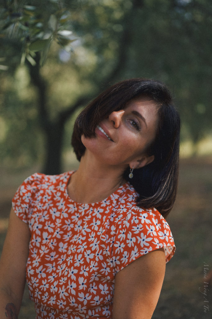 Close-up of a woman with a content smile, looking off to the side, set against a blurred background of leafy trees. She has short dark hair, wears a red dress with a white floral pattern, and simple earrings, conveying a relaxed and joyful demeanor.