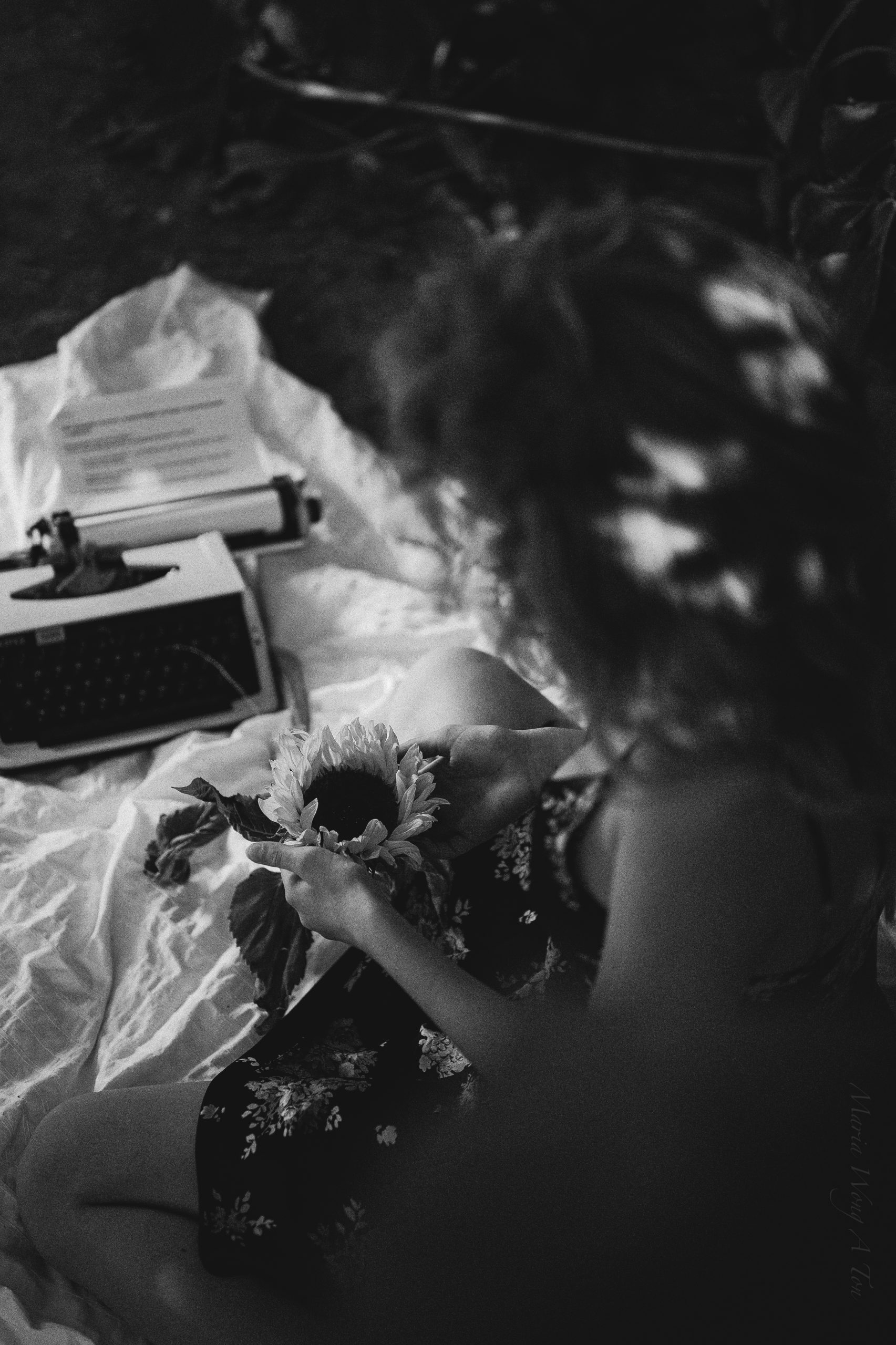 Over-the-shoulder view of a woman in a floral dress holding a sunflower, with a vintage typewriter in the foreground, all set against a moody, dark backdrop, evoking a nostalgic and creative atmosphere