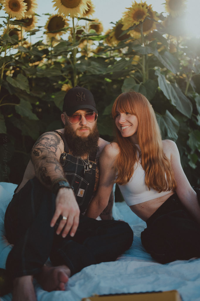 A relaxed couple is nestled together among sunflowers, with the man wearing a cap and showing a tattooed arm around a smiling woman with long red hair, both bathed in soft sunlight.