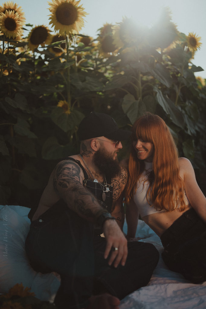 A relaxed couple is nestled together among sunflowers, with the man wearing a cap and showing a tattooed arm around a smiling woman with long red hair, both bathed in soft sunlight.