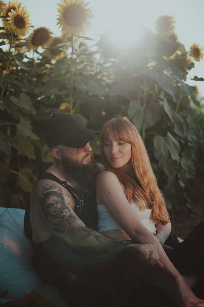 A relaxed couple is nestled together among sunflowers, with the man wearing a cap and showing a tattooed arm around a smiling woman with long red hair, both bathed in soft sunlight.