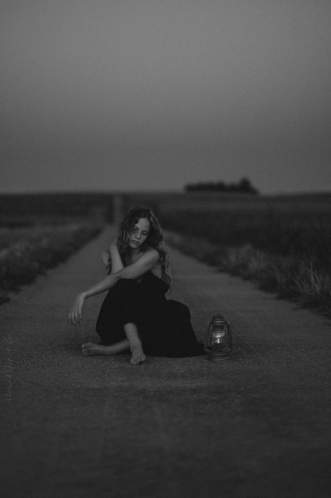 A solemn woman in a black dress sits on a desolate road at dusk, holding a lit lantern, with a distant lighthouse visible in the background, conveying a sense of solitude and contemplation.