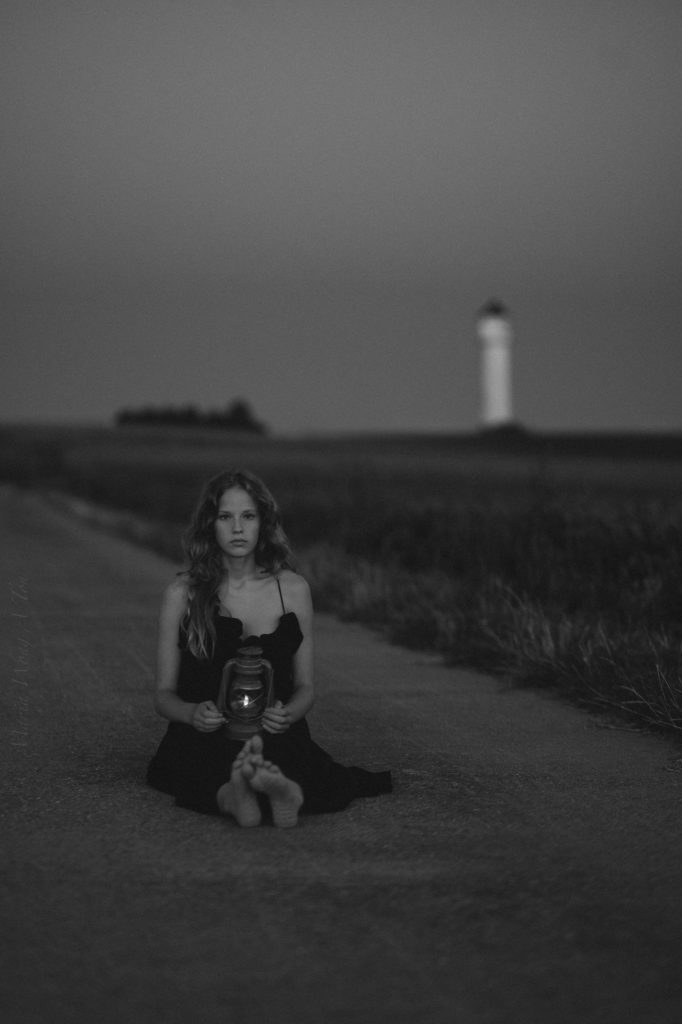 A solemn woman in a black dress sits on a desolate road at dusk, holding a lit lantern, with a distant lighthouse visible in the background, conveying a sense of solitude and contemplation.