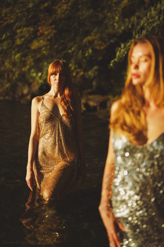 Two women standing in water, one with red hair in a sparkling gold dress in focus and the other blurred in the foreground with a sequined silver dress, both bathed in the golden light of the setting sun filtering through trees.