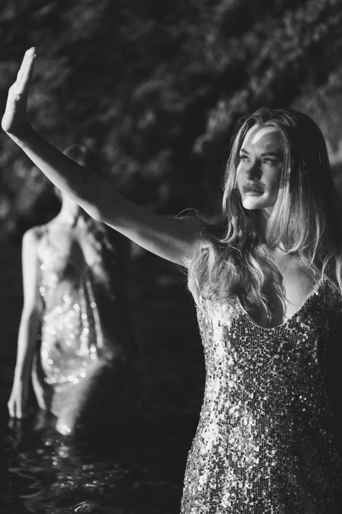 Two women standing in water, one with red hair in a sparkling gold dress in focus and the other blurred in the foreground with a sequined silver dress, both bathed in the golden light of the setting sun filtering through trees.