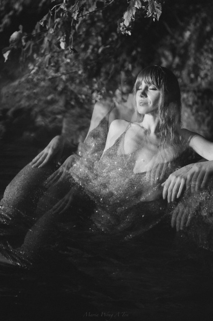 "A joyful woman with red hair, wearing a sparkling gold dress, is seated in water, with sunlight dappling through leaves above, creating a warm, glittering effect as water splashes around her."