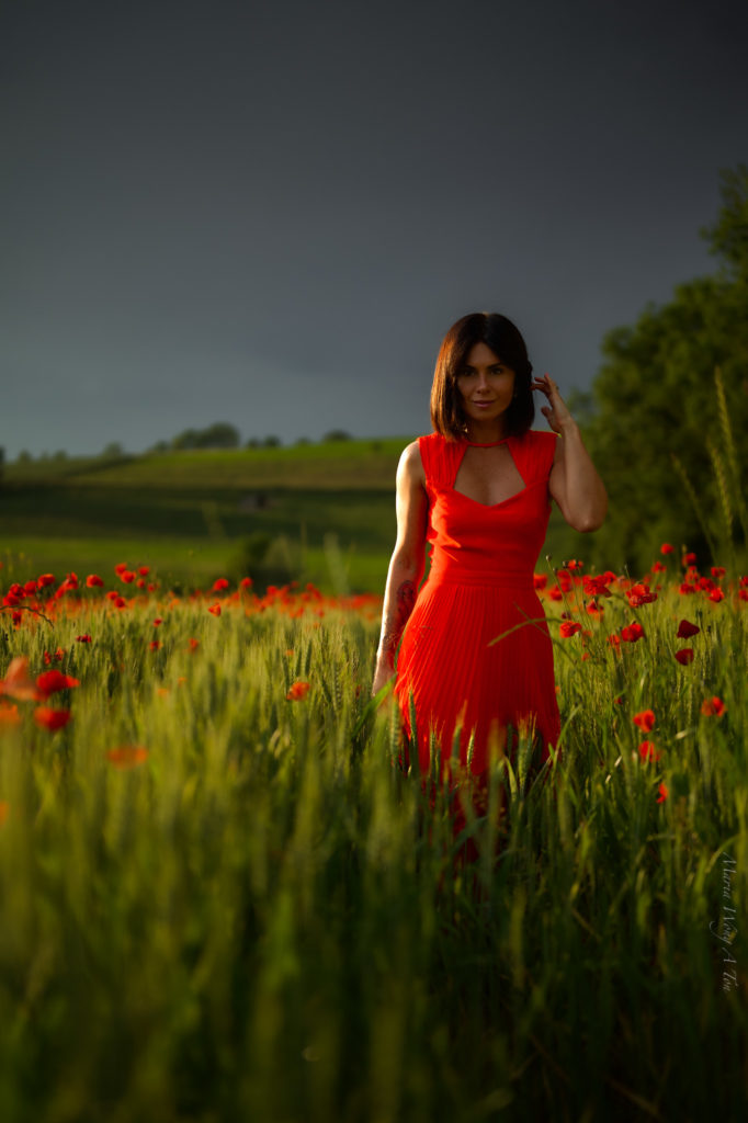 A woman in a vibrant red dress stands amidst a field of green with scattered poppies, her gaze serene and a subtle smile on her lips, with the gentle light of a setting or rising sun casting a warm glow on the scene.