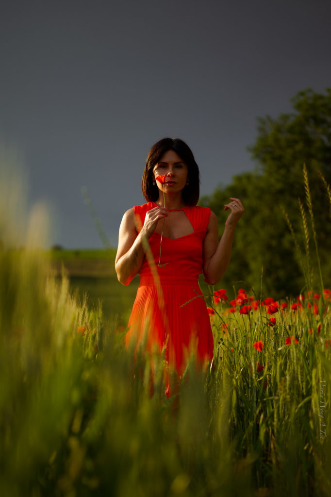 A woman in a vibrant red dress stands amidst a field of green with scattered poppies, her gaze serene and a subtle smile on her lips, with the gentle light of a setting or rising sun casting a warm glow on the scene.