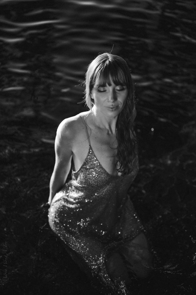 "A joyful woman with red hair, wearing a sparkling gold dress, is seated in water, with sunlight dappling through leaves above, creating a warm, glittering effect as water splashes around her."