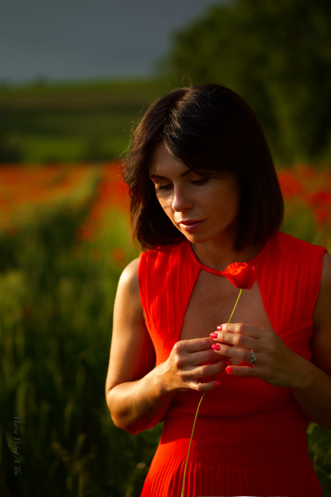 A woman in a vibrant red dress stands amidst a field of green with scattered poppies, her gaze serene and a subtle smile on her lips, with the gentle light of a setting or rising sun casting a warm glow on the scene.