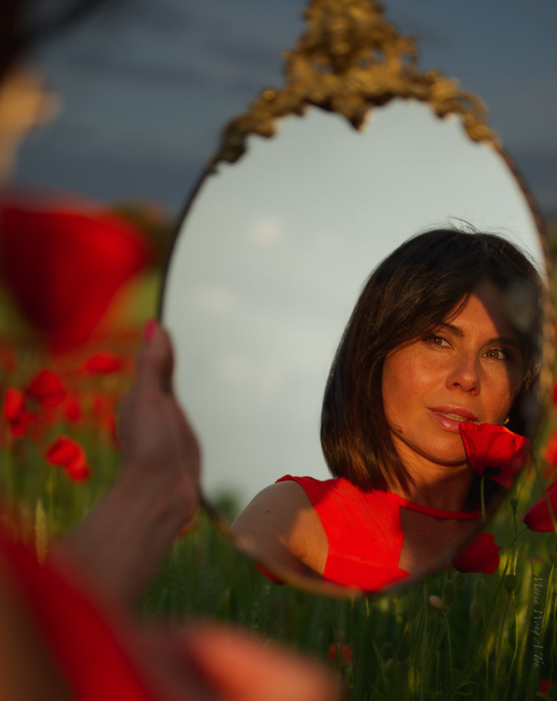 A woman in a vibrant red dress stands amidst a field of green with scattered poppies, her gaze serene and a subtle smile on her lips, with the gentle light of a setting or rising sun casting a warm glow on the scene.