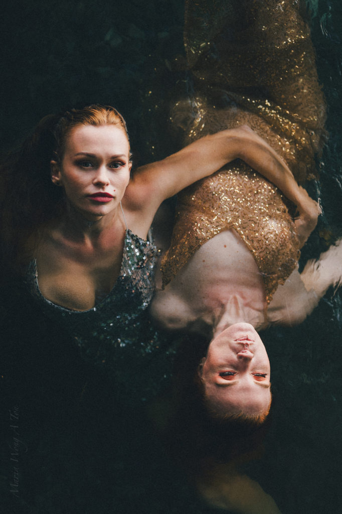 Two women in embellished dresses, partially submerged in water, gaze intently at the camera with a backdrop of shadowy water reflections, creating a moody and evocative portrait.