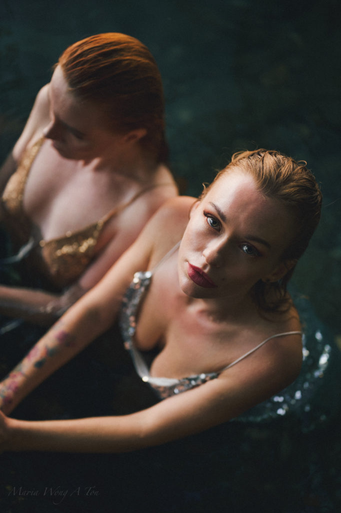 Two women in embellished dresses, partially submerged in water, gaze intently at the camera with a backdrop of shadowy water reflections, creating a moody and evocative black and white portrait.