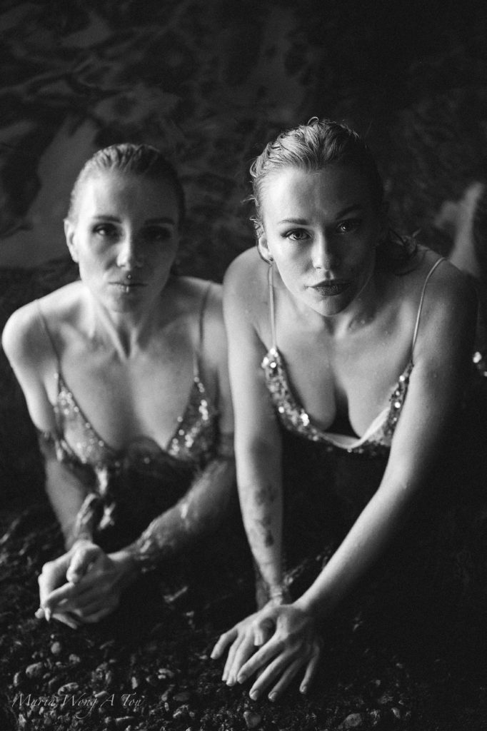 Two women in embellished dresses, partially submerged in water, gaze intently at the camera with a backdrop of shadowy water reflections, creating a moody and evocative black and white portrait.