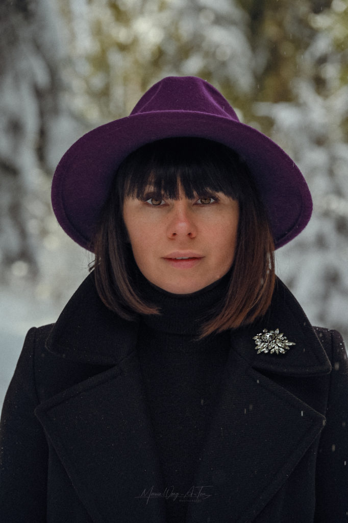 Elegant woman with a contemplative expression wearing a deep purple hat and a black coat with a sparkling brooch, standing against a blurred winter backdrop of snowy trees.