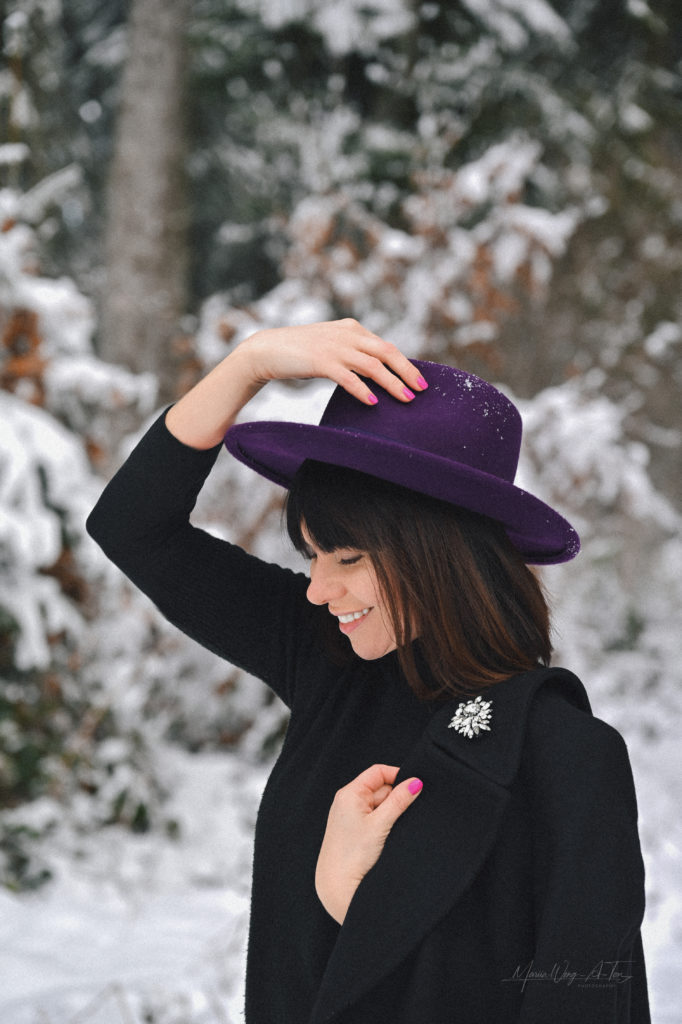 Elegant woman with a contemplative expression wearing a deep purple hat and a black coat with a sparkling brooch, standing against a blurred winter backdrop of snowy trees.