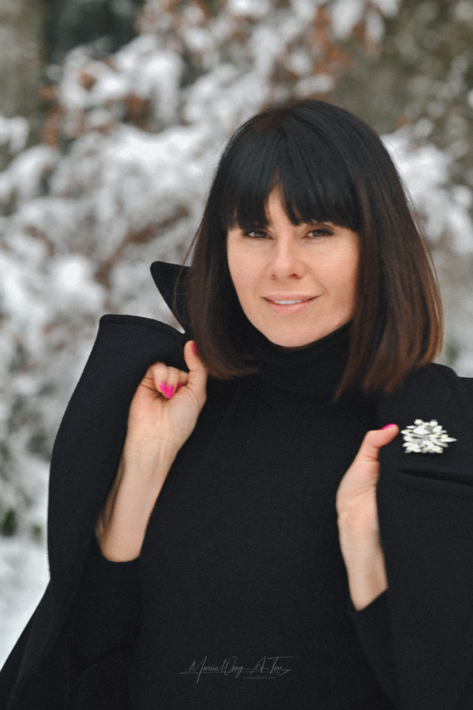 Elegant woman with a contemplative expression wearing a black coat with a sparkling brooch, standing against a blurred winter backdrop of snowy trees.