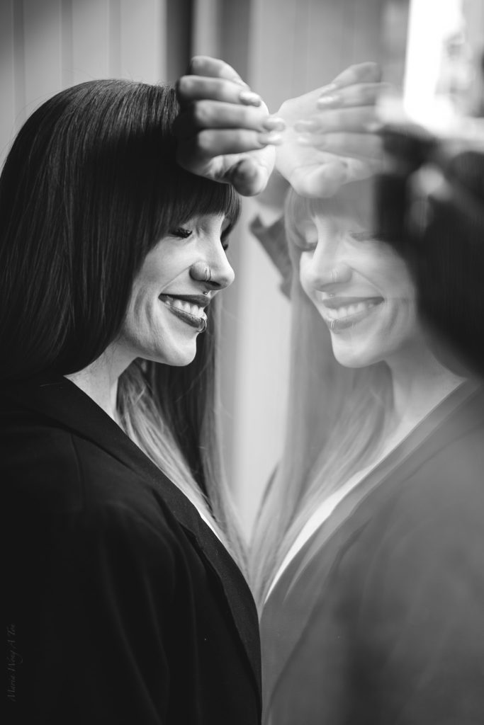 A woman with vivid red hair and a black blazer stands in profile, her face partially reflected in a window, creating a layered image that suggests introspection and self-examination.