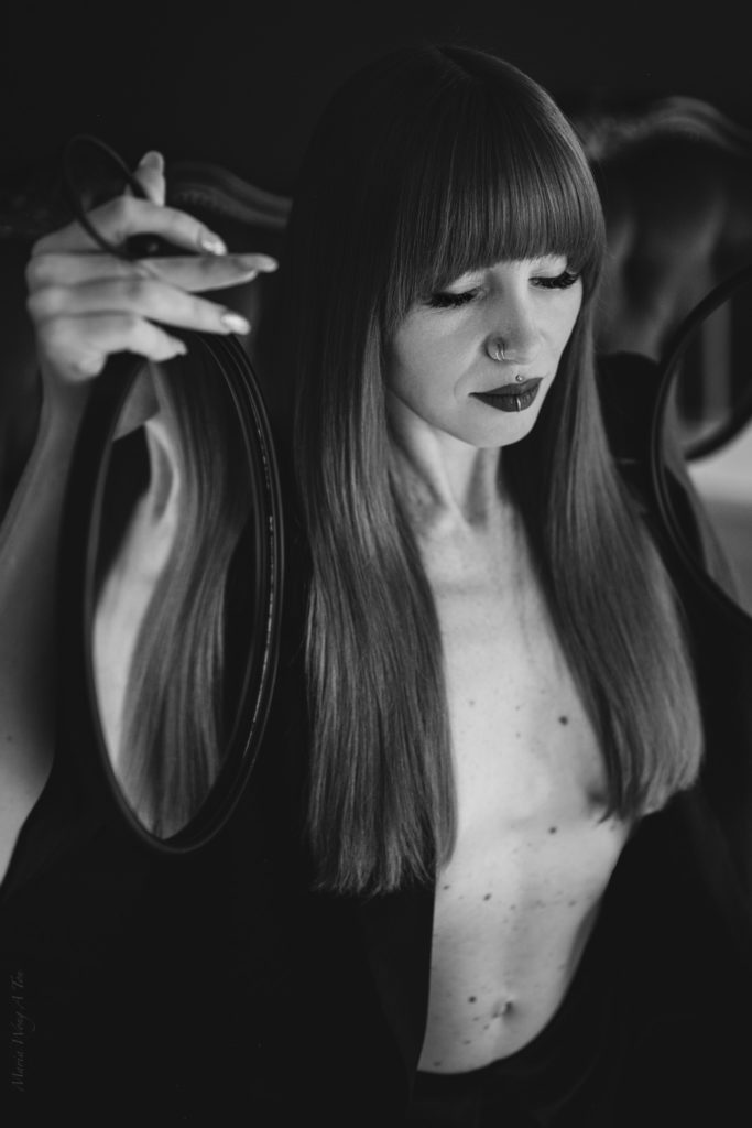 A monochrome image of a woman with straight, long red hair, gazing thoughtfully at her reflection in a handheld mirror, her expression contemplative, set against a dark background that adds to the introspective mood.