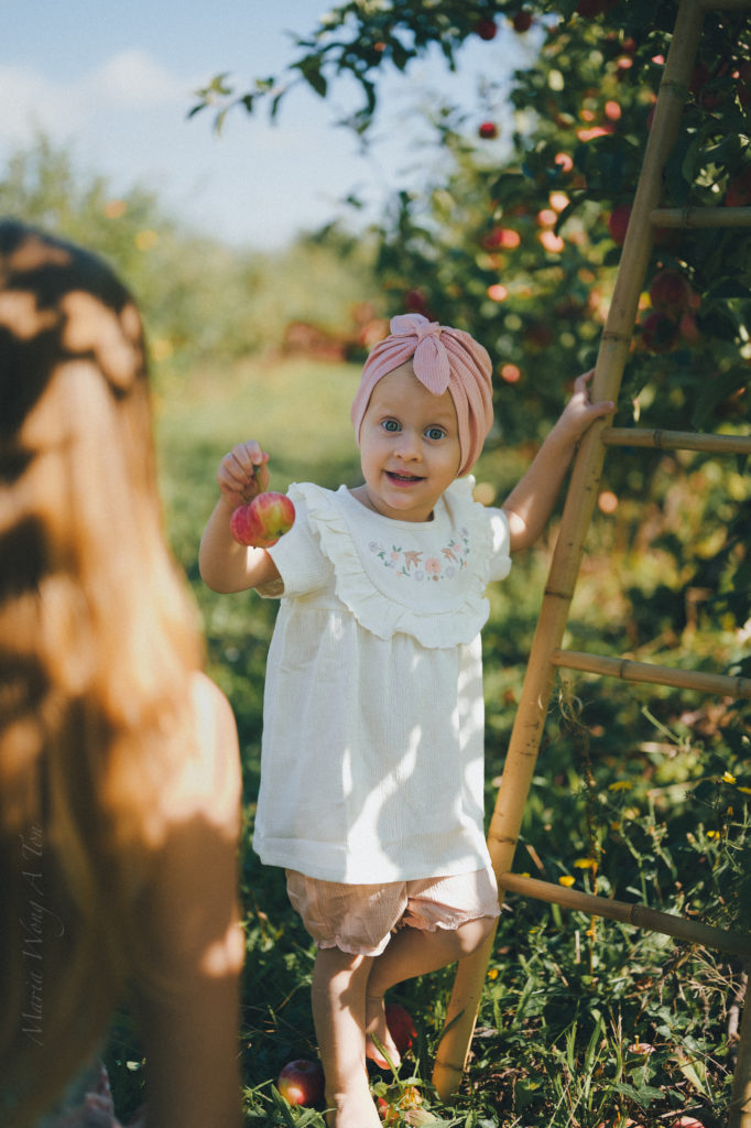 A delighted toddler in a white blouse and pink headband stands in an orchard, offering an apple with a bright, curious expression, as a woman is seen from behind in the foreground, creating a playful and affectionate scene.