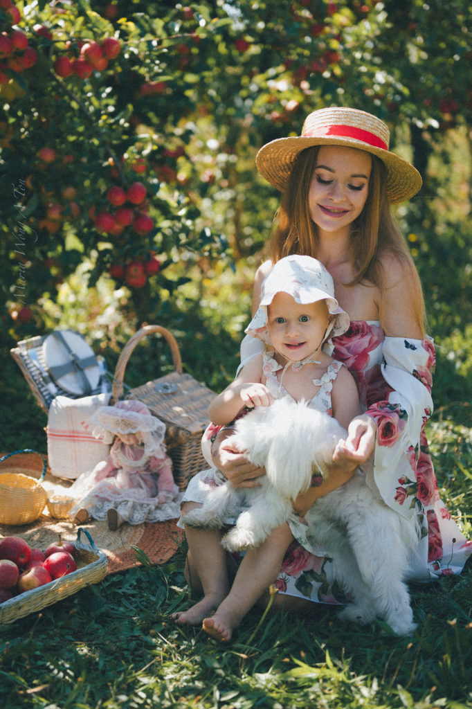 A smiling young woman in a floral dress and a straw hat holds a small white dog and a kid in her arms, seated in a lush garden with roses and foliage in the background, evoking a sense of summertime joy.