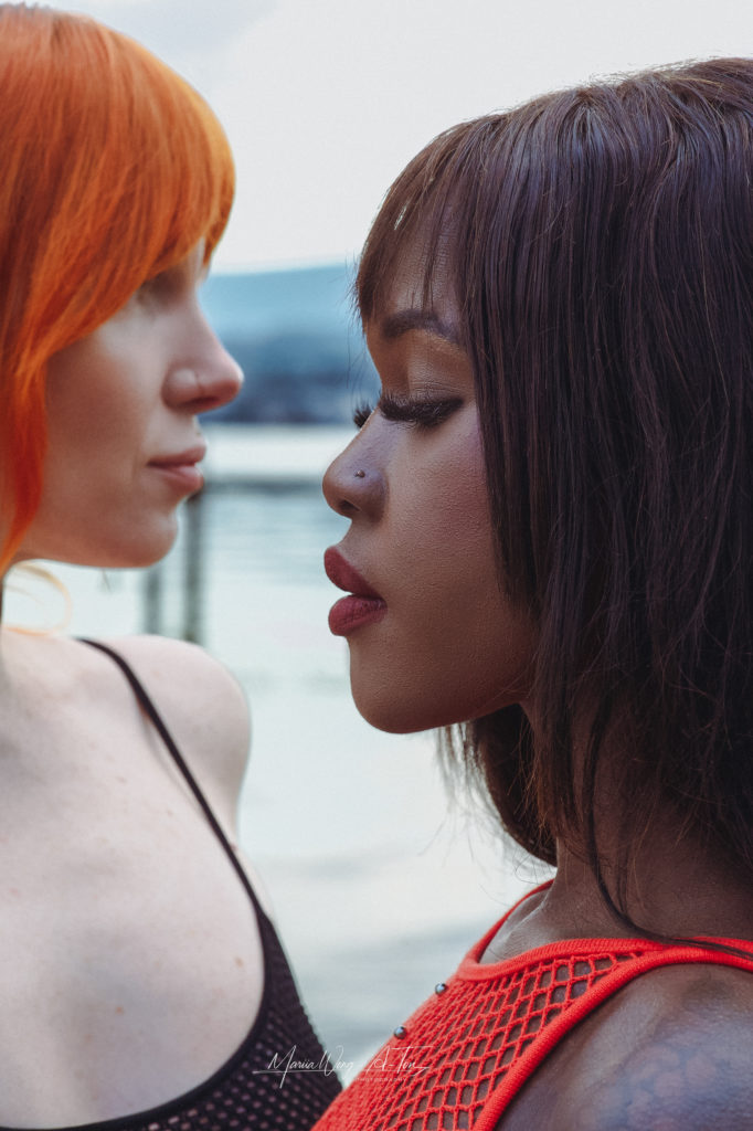 Side profile of two women close together, one with vivid orange hair and the other with dark brown hair, both showcasing a moment of silent communication and connection against a blurred background.