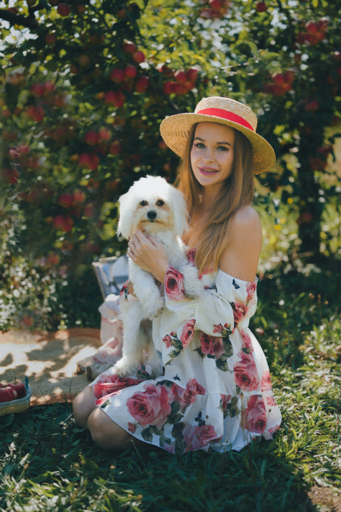 A smiling young woman in a floral dress and a straw hat holds a small white dog in her arms, seated in a lush garden with roses and foliage in the background, evoking a sense of summertime joy.