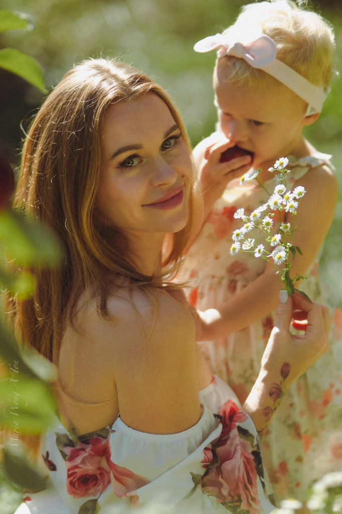 A tender moment, where a smiling woman is adoringly looking at a young child who is holding a small bouquet of flowers. The child, wearing a floral dress and a headband, leans affectionately against the woman in a sunlit outdoor setting.