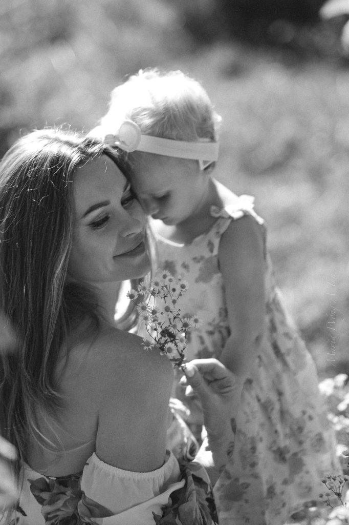 A tender moment captured in black and white, where a smiling woman is adoringly looking at a young child who is holding a small bouquet of flowers. The child, wearing a floral dress and a headband, leans affectionately against the woman in a sunlit outdoor setting.