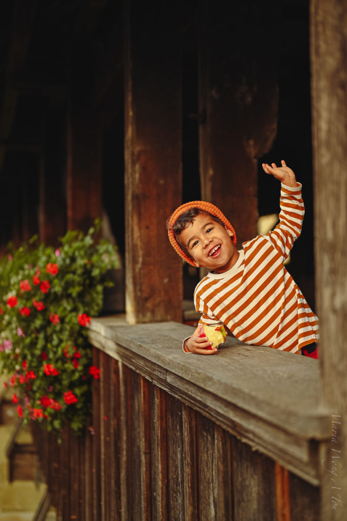 A cheerful young child in a striped orange and white shirt and a knitted hat, playfully waving and holding an apple, peeks over a rustic wooden balcony adorned with vibrant red flowers.