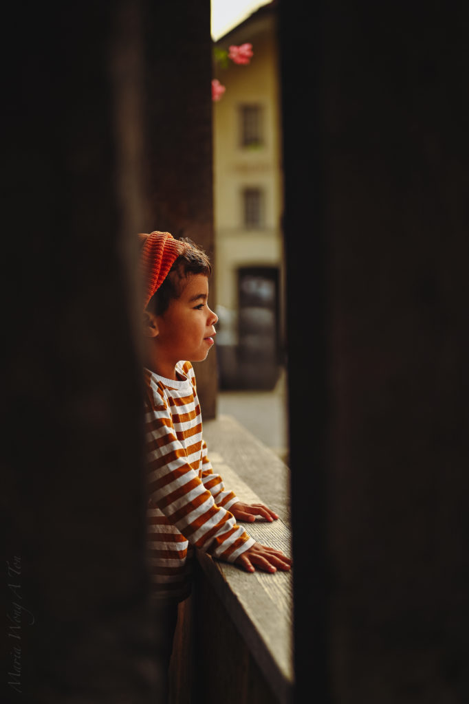 A cheerful young child in a striped orange and white shirt and a knitted hat, peeks over a rustic wooden balcony adorned with vibrant red flowers.