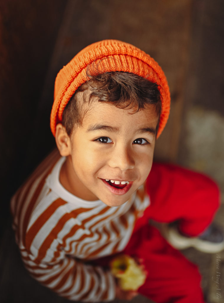 A cheerful young child in a striped orange and white shirt and a knitted hat, peeks over a rustic wooden balcony adorned with vibrant red flowers.