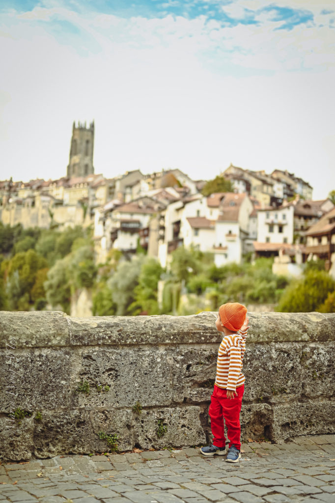 A small child in a striped orange and white top and red pants stands by a stone parapet, gazing at a picturesque town with historic buildings and a prominent tower in the soft-focus background, under a cloudy sky.