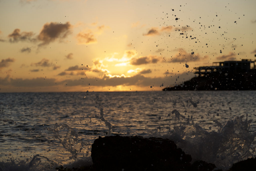 Sunset over the ocean, with the sun's golden light piercing through scattered clouds above the horizon, and water droplets captured in mid-air from a wave crashing against the rocks.