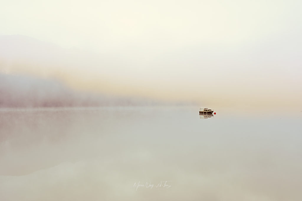 A solitary boat floats on a tranquil lake enveloped in mist, with soft pastel colors of dawn creating a serene and almost ethereal landscape.