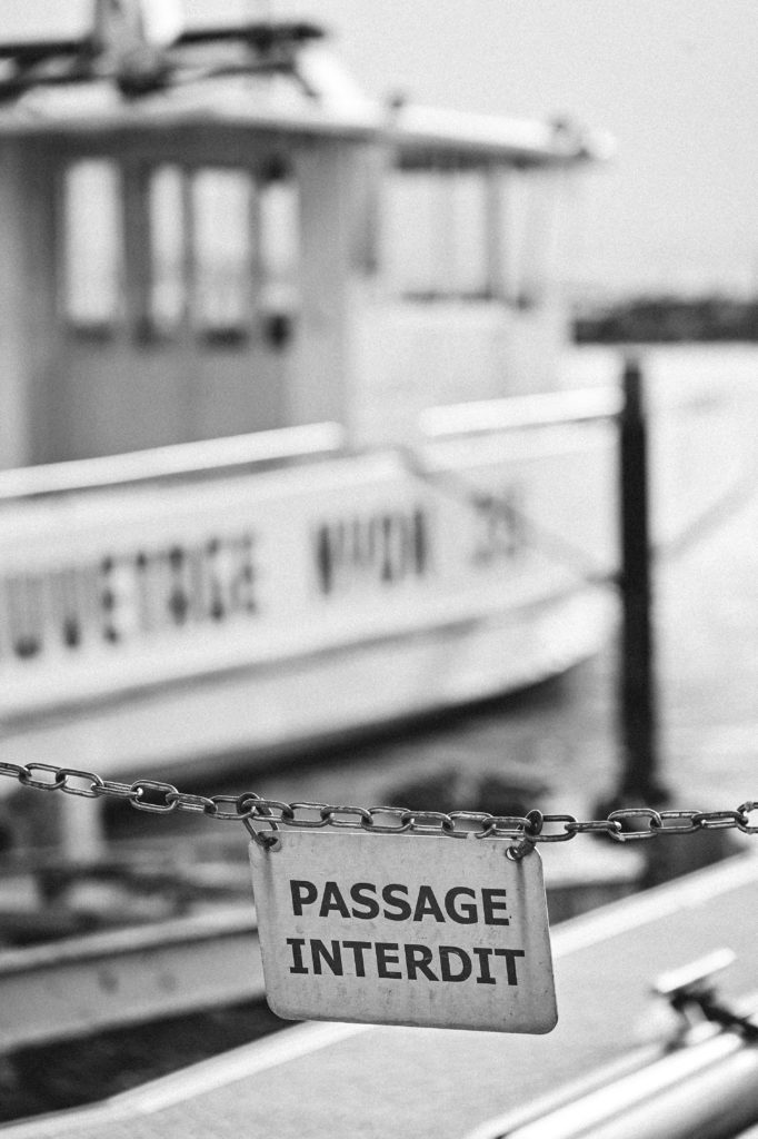 Black and white image focused on a 'PASSAGE INTERDIT' sign hanging on a chain with a blurred boat named 'MONTREUX No. 75' in the background, symbolizing restricted access at a dock
