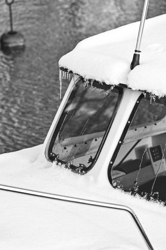 Black and white image displaying a close-up of a boat's snow-covered canopy, with icicles hanging from its edge, set against a blurry background of water.