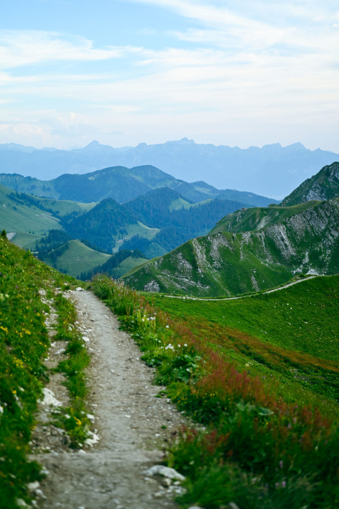 An idyllic mountain ridge with a narrow trail at sunset, overlooking a vast, verdant landscape with layers of hills stretching into the distance under a soft pink and blue sky.