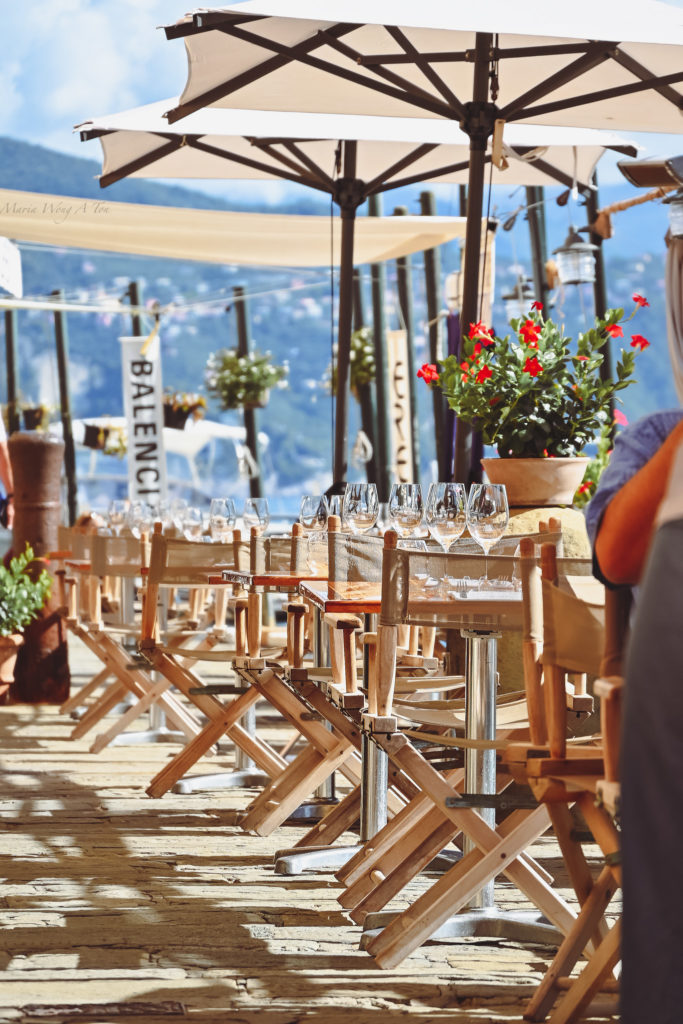 An inviting outdoor cafe setting with neatly arranged wooden chairs and sparkling wine glasses laid out on tables, vibrant red flowers adding a pop of color, all under the shade of large beige umbrellas, with a view of a mountainous landscape in the background, suggesting a leisurely dining experience in a scenic location.