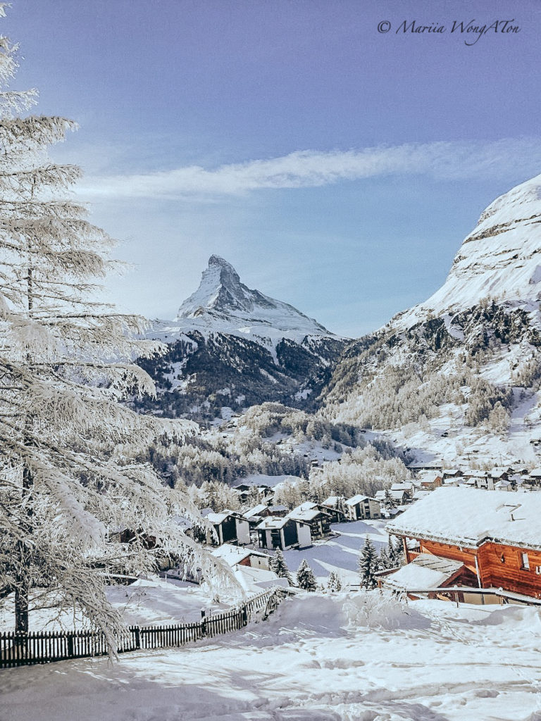 A picturesque winter scene capturing the majestic Matterhorn mountain towering over a quaint village. The landscape is blanketed in snow, reflecting a serene and crisp winter day, with snow-laden trees and traditional wooden chalets, creating a quintessential alpine postcard image.
